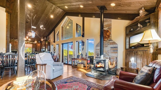 A spacious living room with a fireplace, wooden beams, and a large window overlooking a scenic view. - The Barn at Quail Creek Ranch - 2 Bedroom Peaceful Ranch Retreat (Wheeler)