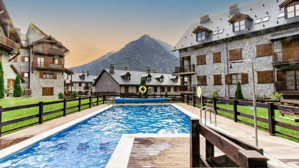 Un hotel con piscina, edificios de piedra y una montaña al fondo.