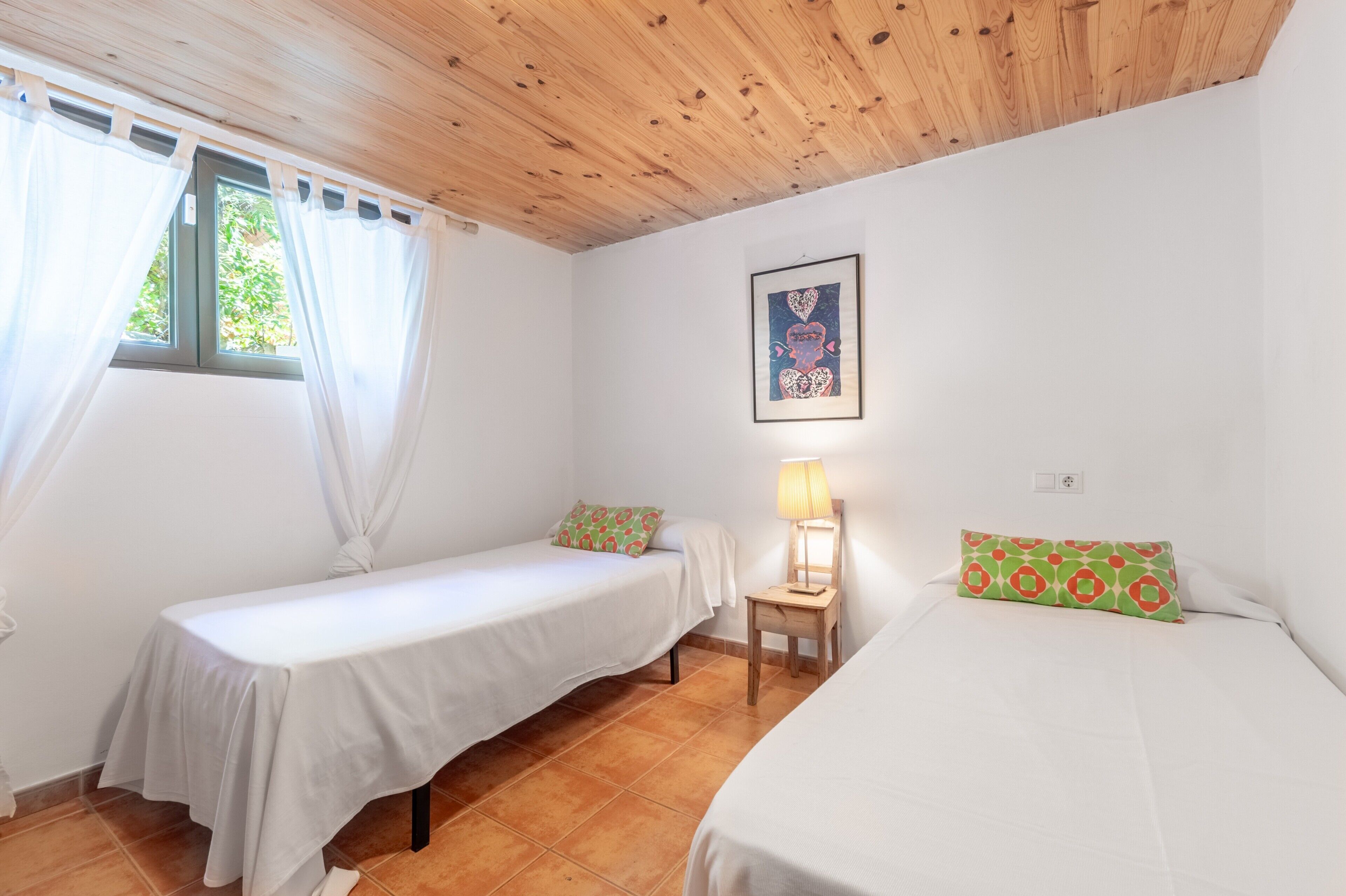 A room with two single beds, a small table with a lamp, a window with curtains, and a framed picture on the wall.