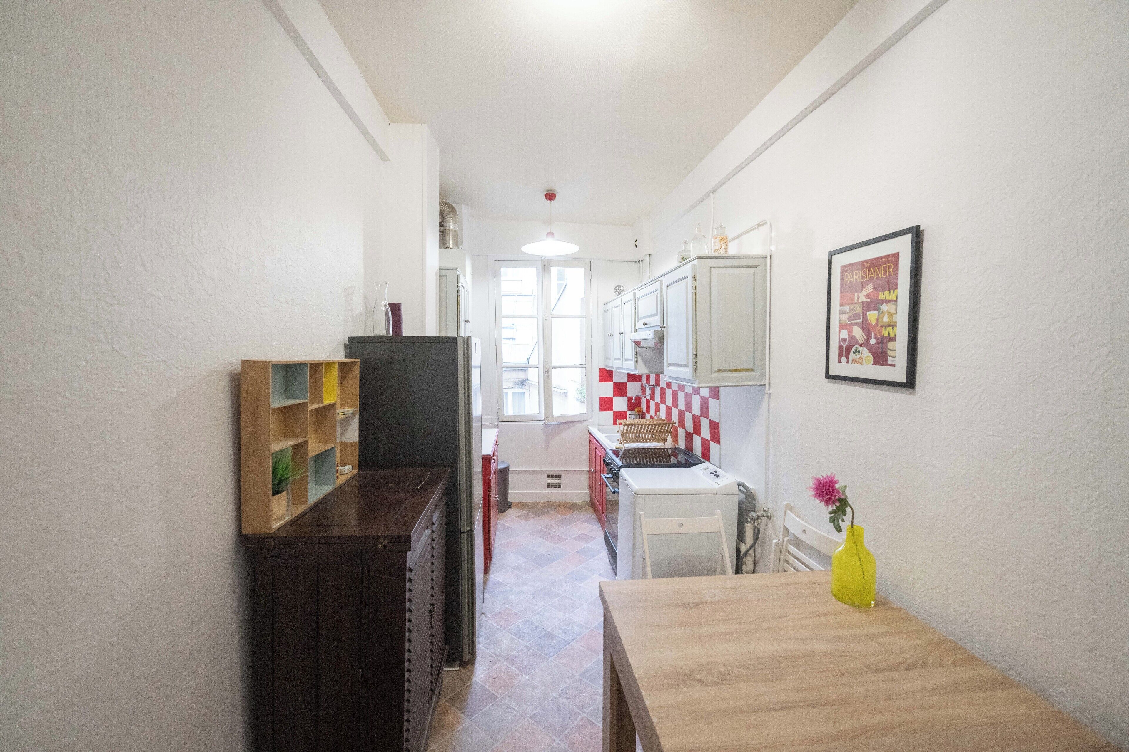A narrow hallway with a wooden table and a yellow vase on the right, leading to a kitchen area with a red and white checkered backsplash and cabinets.