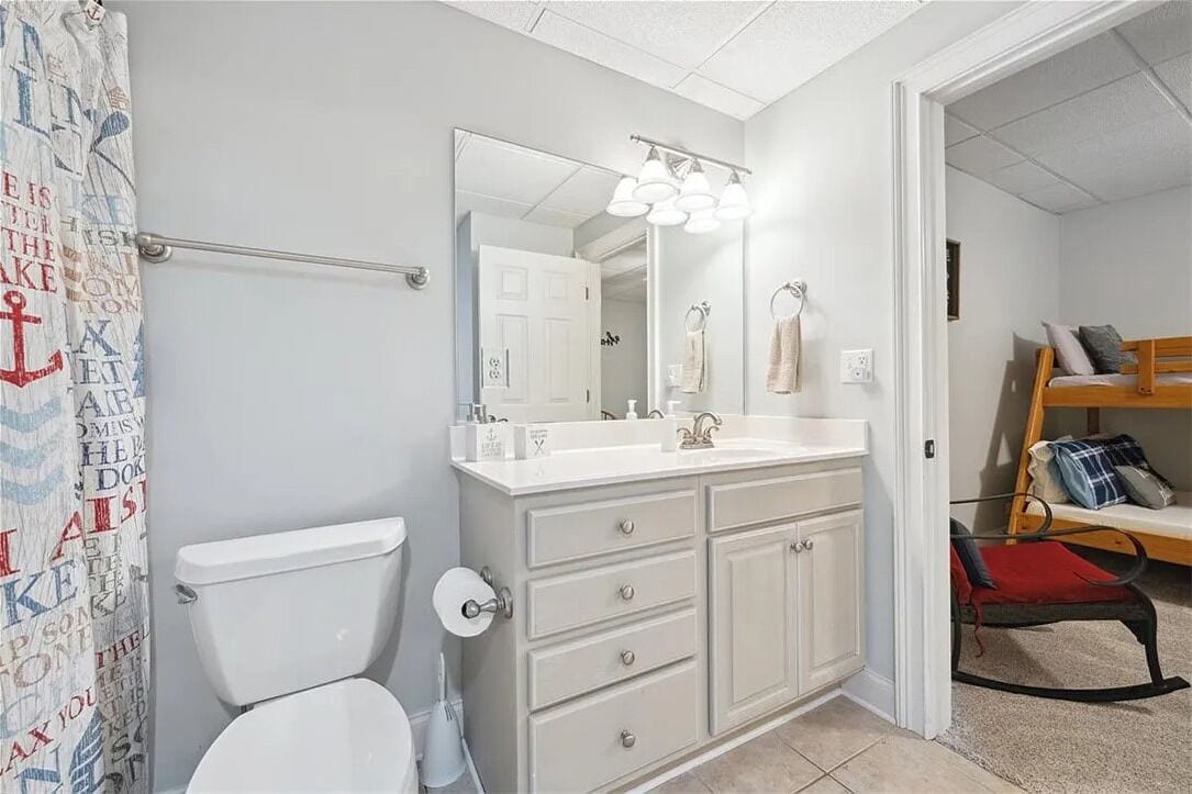 Une salle de bain comprenant des toilettes, un meuble-lavabo avec un miroir et des luminaires, et un rideau de douche orné d’un motif nautique.