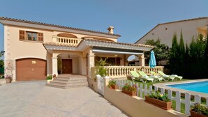 Una casa de dos pisos con piscina, un porche cubierto con sillones de descanso y una entrada con portón.