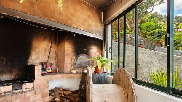 A brick fireplace with a log storage area, a potted plant, and a view of a hillside through a window. - Traveller's House Holiday Home with Mountain View, Shared Pool, and Wi-Fi (Madeira Machico)