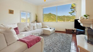 A living room with a sofa, a fireplace, a large window with a mountain view, and a television. - Traveller's House Holiday Home with Mountain View, Shared Pool, and Wi-Fi (Madeira Machico)