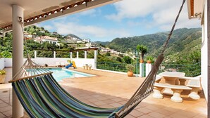 A hammock, a pool, and a view of houses on a hillside. - Traveller's House Holiday Home with Mountain View, Shared Pool, and Wi-Fi (Madeira Machico)