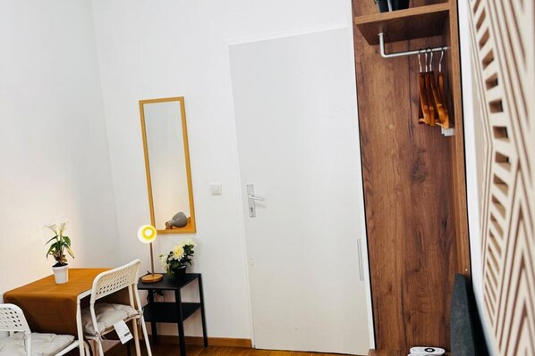 A room with a wooden door, a white wall, a mirror, a desk with a chair, and a small table with a lamp.