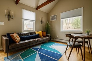 Ein Wohnzimmer mit einem braunen Ledersofa, einem blau-weißen Teppich, einem Esstisch mit Stühlen und zwei Fenster mit Jalousien.