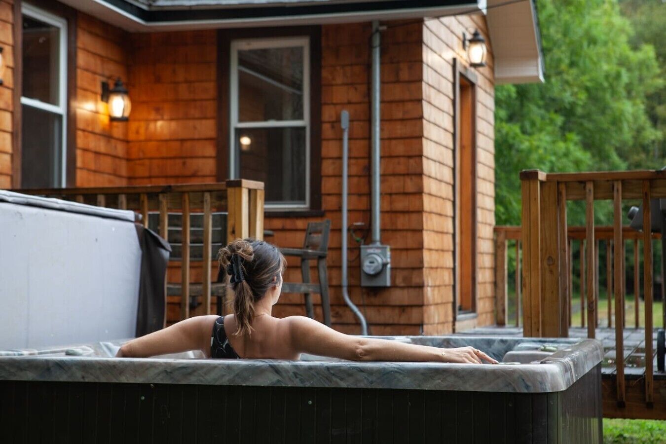 Une personne se détend dans un bain à remous situé à l’extérieur d’un chalet en bois, qui possède une terrasse et du mobilier d’extérieur.
