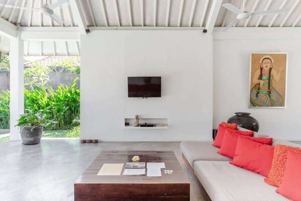 A modern living room with a flat-screen TV mounted on the wall, a wooden coffee table with papers, a white sofa with red cushions, and a large vase.