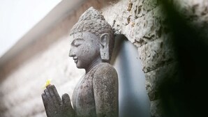 En sten Buddhastatue med en hånd hævet, placeret foran en ru stenvæg.