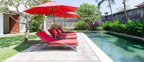 Un espace aménagé au bord de la piscine, avec des chaises longues rouges et un parasol rouge, entouré de verdure et donnant sur une piscine aux eaux cristallines.