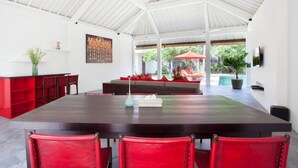 En rummelig spiseafdeling med et langt bord og røde stole, hvidt loft samt udsigt til en udendørs terrasse.