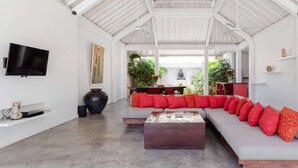 A spacious living area with a high ceiling, a flat-screen TV mounted on the wall, a large dark vase, and a long sofa with red cushions.
