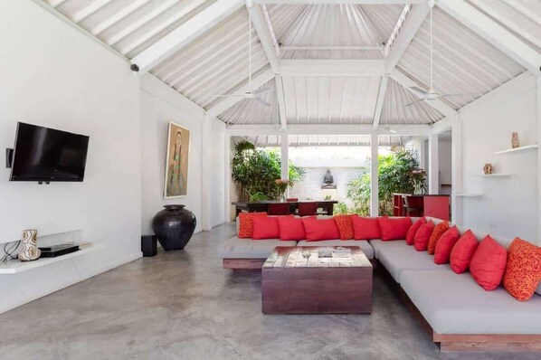 A spacious living area with a high ceiling, a flat-screen TV mounted on the wall, a large dark vase, and a long sofa with red cushions.