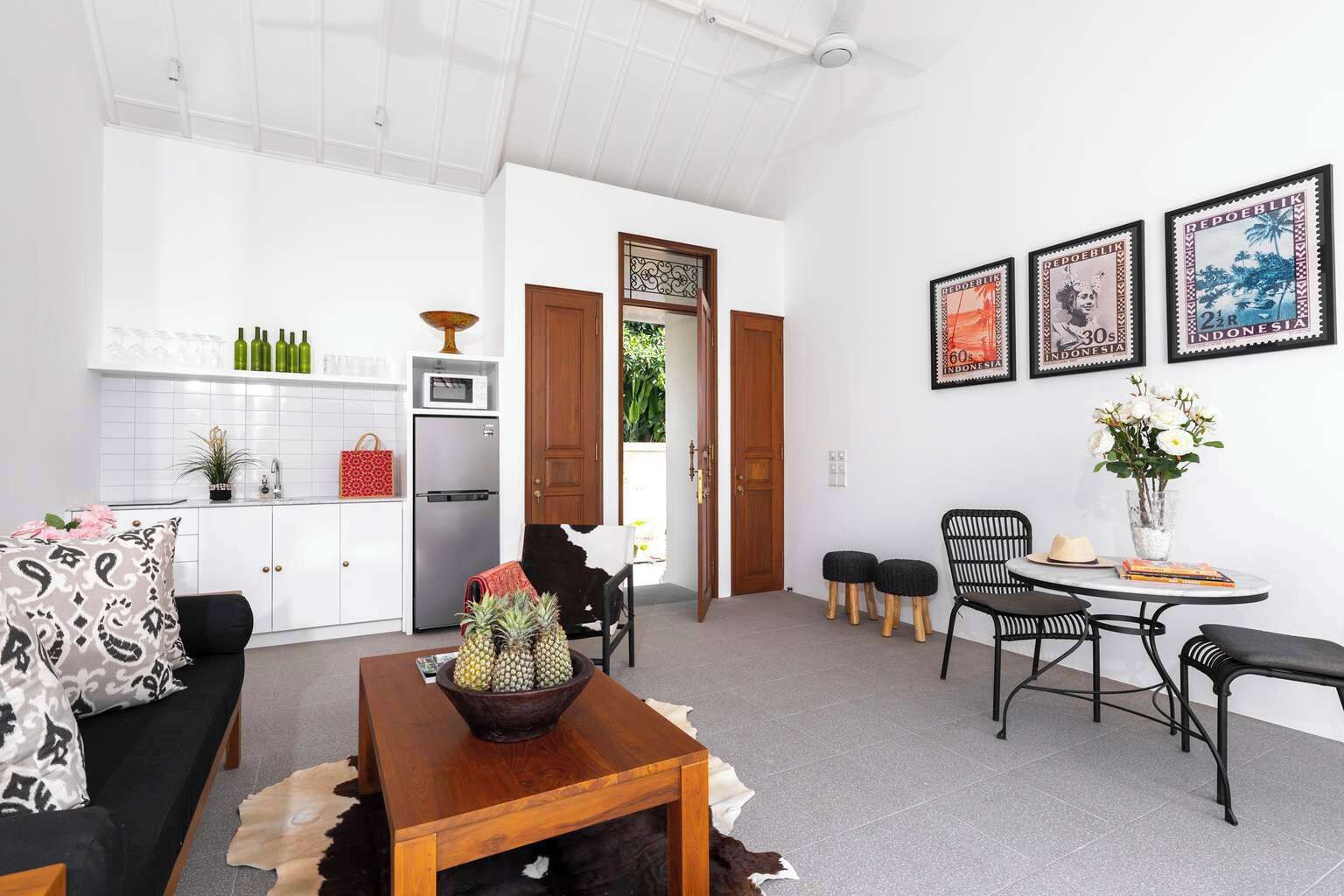 A modern living room with a sofa, coffee table, and a dining area with chairs and a table. There is a kitchen area with a refrigerator, microwave, and a cabinet. The room has white walls and a ceiling fan.