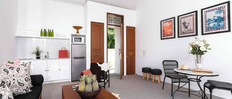 A modern living room with a sofa, coffee table, and a dining area with chairs and a table. There is a kitchen area with a refrigerator, microwave, and a cabinet. The room has white walls and a ceiling fan.