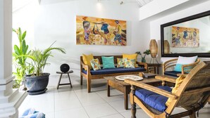 Ein geräumiger Wohnbereich mit einer Holzsofa, einem Korbstuhl und einem Couchtisch. An der Wand befinden sich zwei große Gemälde, ein Blumentopf und ein kleiner Beistelltisch.