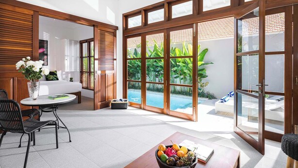 A modern hotel room with a large sliding glass door leading to a pool area, a dining area with a table and chairs, and a view of lush greenery. - Chic Villa Bella | Steps from Eat Street Seminyak (Seminyak)