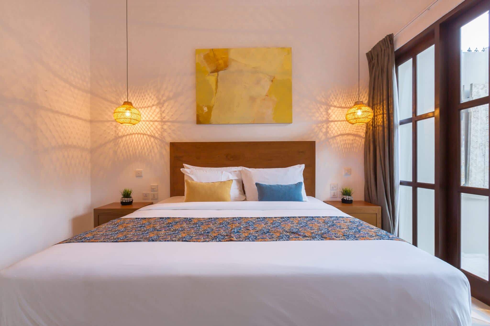 A neatly made bed with a patterned bedspread, two bedside tables with small plants, a wooden headboard, and a large window with curtains.
