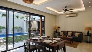A modern living room with a dining area, large windows, and a ceiling fan. - Closed Living Room Villa The Cabana in Seminyak (Seminyak)