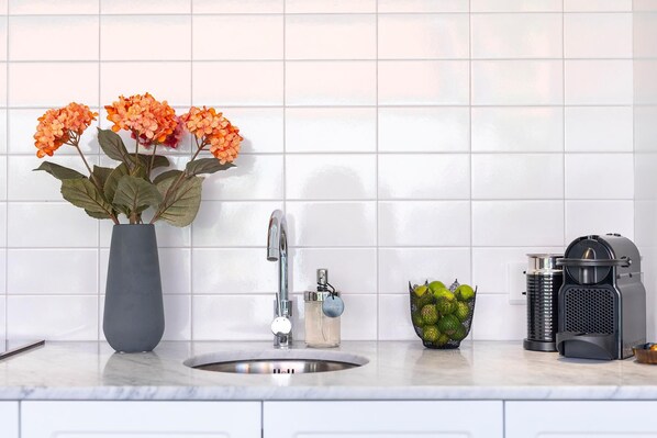 Une cuisine avec un mur recouvert de carreaux blancs, un évier, un robinet, un vase avec des fleurs, un petit bol de citrons et une machine à café.