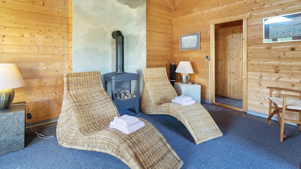Interior de una cabaña de madera con dos sillones de mimbre, un estufa de leña y un cuadro en la pared.