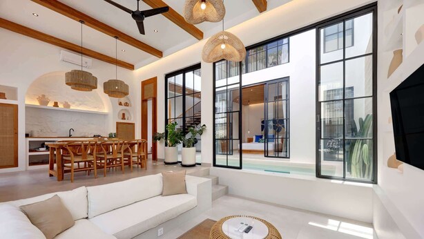 A modern living room with a white sofa, a round wicker coffee table, and large windows offering views of the outdoors.