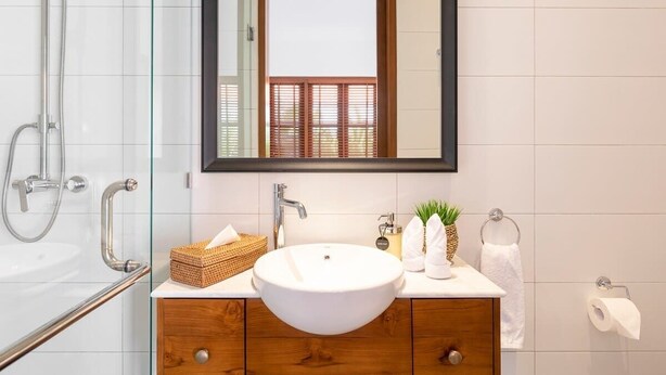 A bathroom with a glass shower enclosure, a white sink, wooden cabinets, and a mirror reflecting a window with blinds. - Cute 1BR Villa 5Mins Walk to Eat Street Seminyak (Seminyak)
