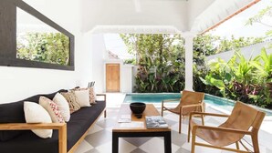 A modern living room with a black sofa, wooden coffee table, and wicker chairs. A pool is visible in the background, surrounded by lush greenery.