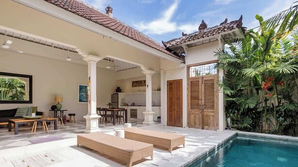 A poolside area with lounge chairs, a kitchen in the background, and tropical plants.