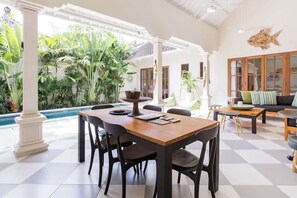 A spacious indoor area with a dining table, chairs, a sofa, and a large window overlooking a pool and lush greenery. - Unwind at Seminyak Pool Villa 1Min Walk to Center (Kerobokan Kelod)