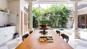 A dining area with a long wooden table, surrounded by chairs, in a room with white walls and a tiled floor.