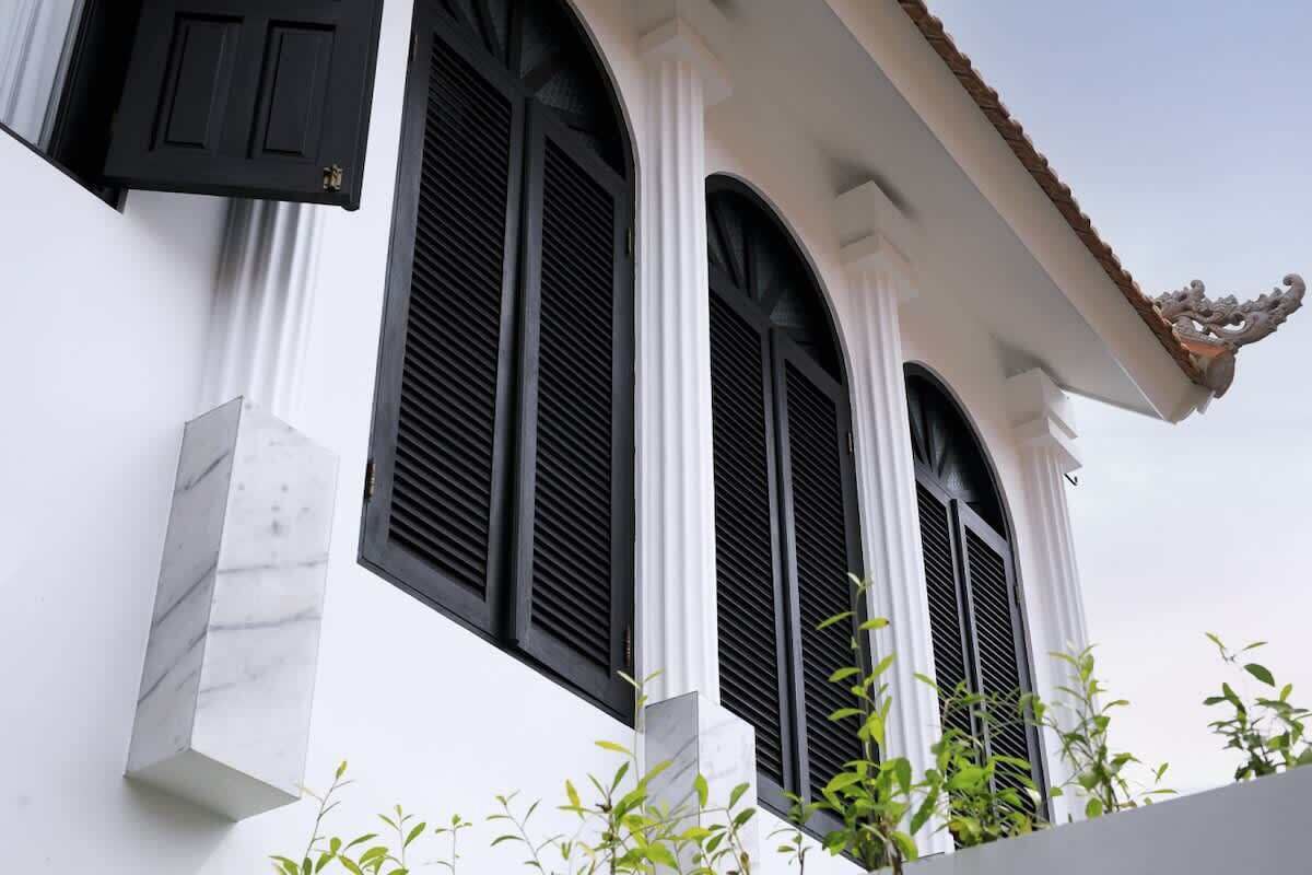 Un bâtiment aux murs blancs et aux volets noirs, présentant un style architectural traditionnel avec des fenêtres en arc de cercle et un élément décoratif sur le toit.