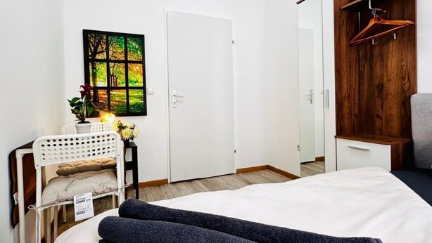 A bedroom with a bed, a white wardrobe, a wooden wardrobe, a white chair, a small table with a vase and flowers, and a painting on the wall. - Affordable Room in Shared Apt. Close to Stadthalle (Vienna)