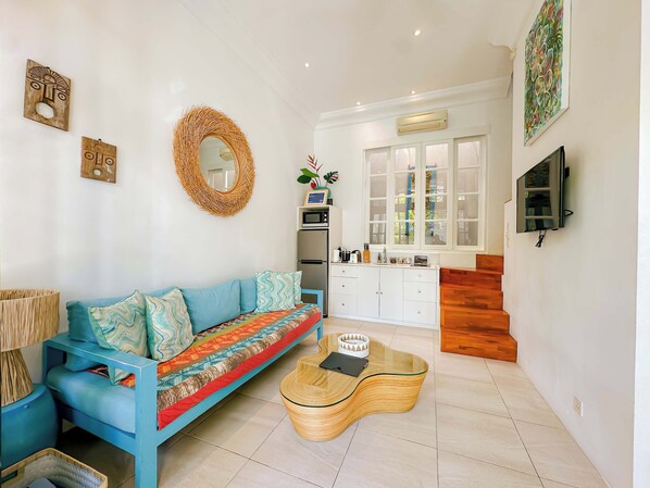 A living room with a blue sofa, a woven mirror, a kitchen with a microwave, and a TV mounted on the wall.