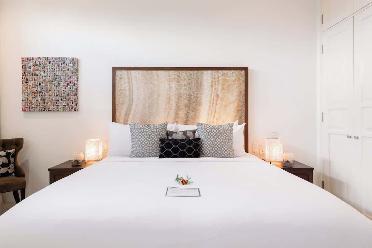 Una cama bien hecha con una colcha blanca y dos almohadas, un cuadro abstracto de gran tamaño en la pared, y una mesita con una lámpara y una vela.