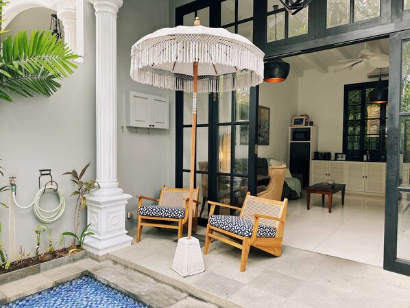 A spacious patio with two wooden chairs, a white umbrella, a small pool, and a view into the interior of the building.