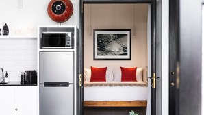 A compact kitchen with a microwave, a coffee machine, and a small refrigerator. There is a bed with red and white bedding, a framed picture on the wall, and a potted plant on a table. - Villa Saffron – Walk to Seminyak's Best Cafe Shop (Kerobokan Kelod)