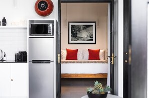 Cocina compacta con microondas, cafetera y refrigerador pequeño. Hay una cama con ropa de cama roja y blanca, un cuadro enmarcado en la pared y una planta en maceta sobre una mesa.