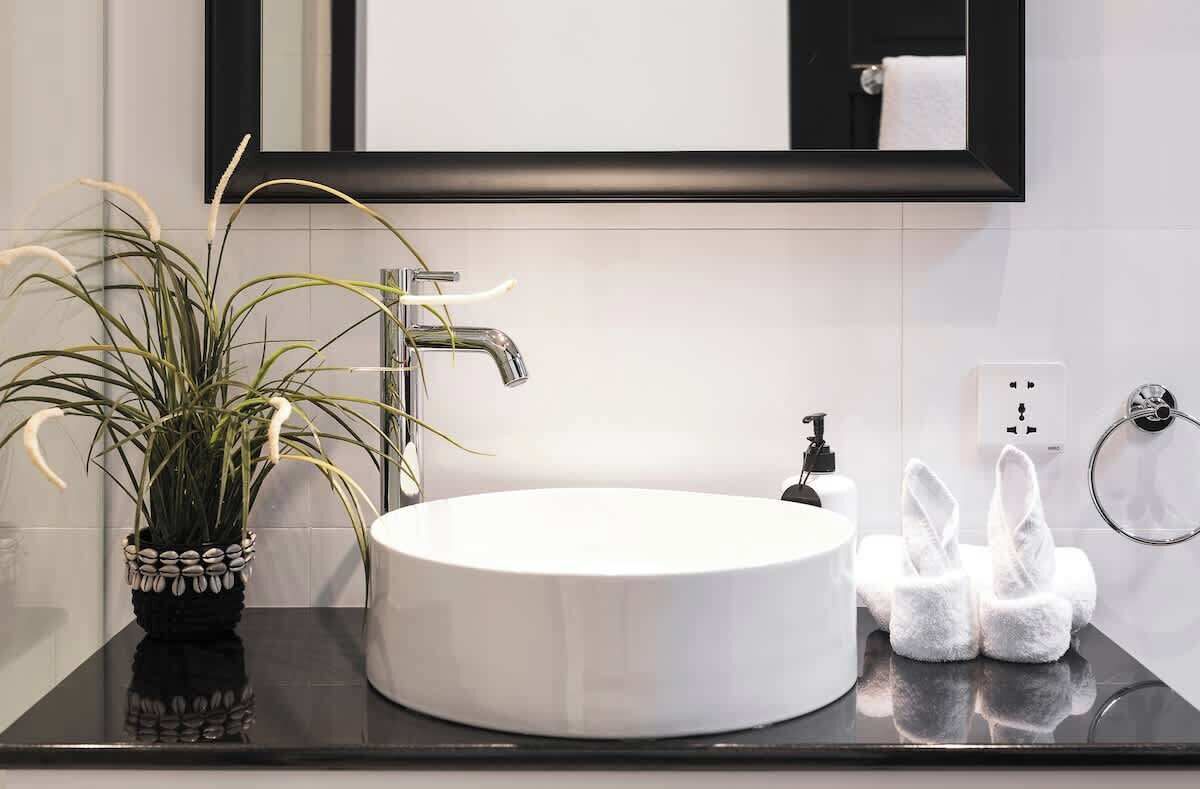 A modern bathroom with a round white sink, a black countertop, a potted plant, a towel rack with white towels, and a mirror above the sink.