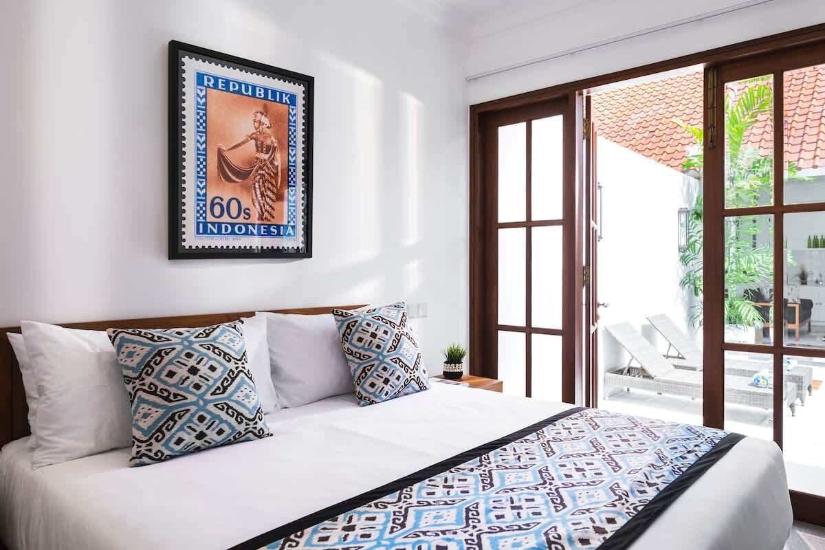 A hotel room with a bed, pillows, a framed Indonesian stamp, and a view through a glass door.