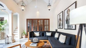 A spacious living room with a dark blue sectional sofa, a round coffee table, and a patterned rug. There are framed artworks on the walls and a large window providing natural light. - Comfort 3BR Soleh Walk to Shops & Beach Seminyak (Kerobokan Kelod)