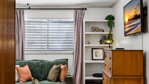 A living room with a green sofa, a wooden cabinet, a TV on the wall, and a window with curtains. - 4 Blocks to Beach! • Historic Cottage • 5mins Strand/Port w Driveway Parking (Galveston)