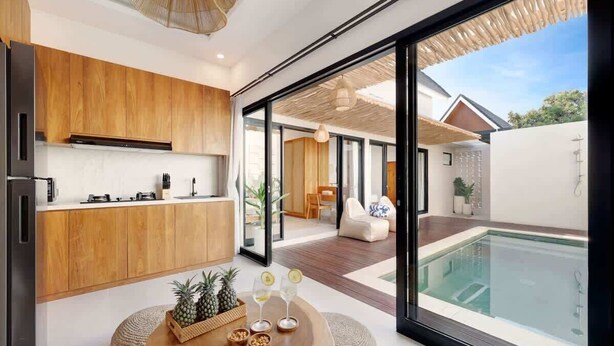 A modern kitchen with wooden cabinets, a dining area with a round wooden table, and a pool visible through large glass doors.