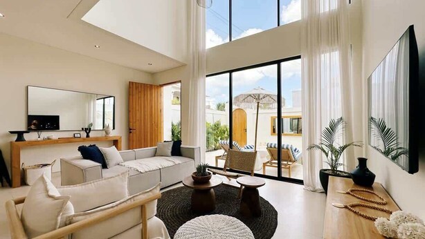 A spacious living room with a sofa, coffee table, and chairs, large windows with curtains, and a flat-screen TV on the wall.