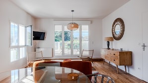 Una sala de estar moderna con una mesa de comedor de vidrio, un sofá, un televisor, una silla de mimbre, un mueble de madera y un ventanal con vistas.