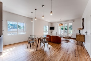 A spacious room with wooden flooring, a dining table with chairs, a sofa, and a television mounted on the wall.