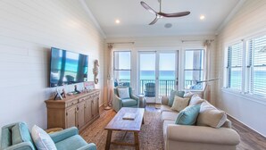 A spacious living room with a large flat-screen TV, comfortable seating, a ceiling fan, and a view of the ocean through large windows. - Journeys End 205 - Welcome to your coastal escape! Gulf Front! Private Beach! (Santa Rosa Beach)