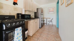 Una cocina compacta con estufa, microondas y refrigerador. Hay una pequeña mesa de comedor con dos sillas. La cocina tiene gabinetes de madera clara y piso de baldosas.