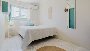 Un dormitorio con una cama, una ventana con cortinas, un aire acondicionado instalado en la pared, un colgador de macramé, una puerta y un taburete pequeño.